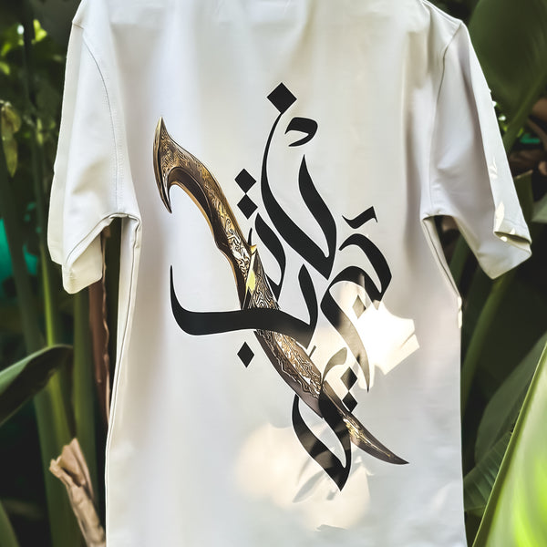 White t-shirt with Arabic calligraphy design on a hanger against a green leafy background