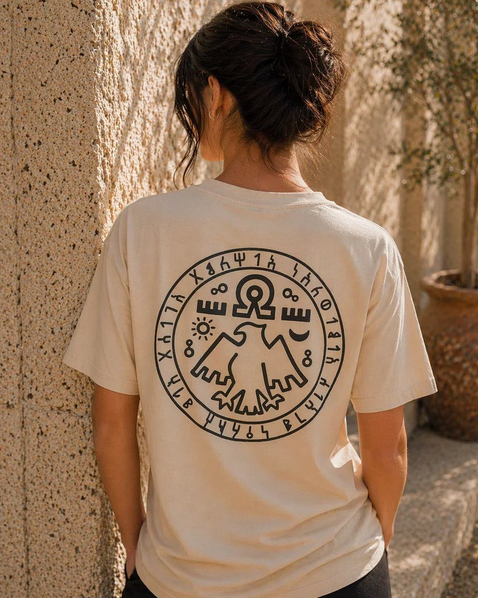 Person wearing a beige t-shirt with a black circular design on the back, standing against a textured wall.
