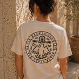 Person wearing a beige t-shirt with a black circular design on the back, standing against a textured wall.