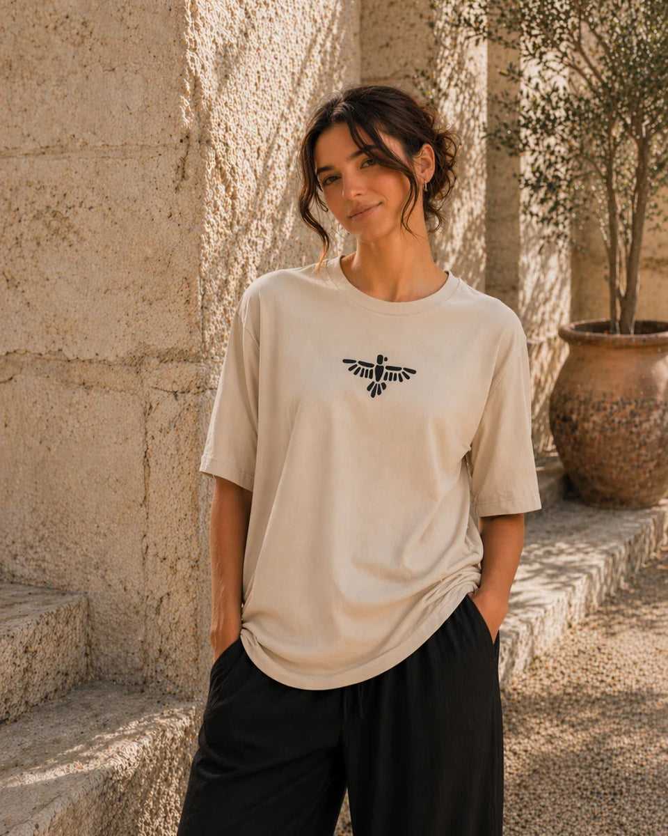 Woman wearing a beige t-shirt with a logo, standing outdoors against a stone wall.