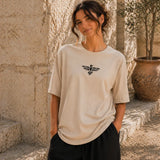 Woman wearing a beige t-shirt with a logo, standing outdoors against a stone wall.