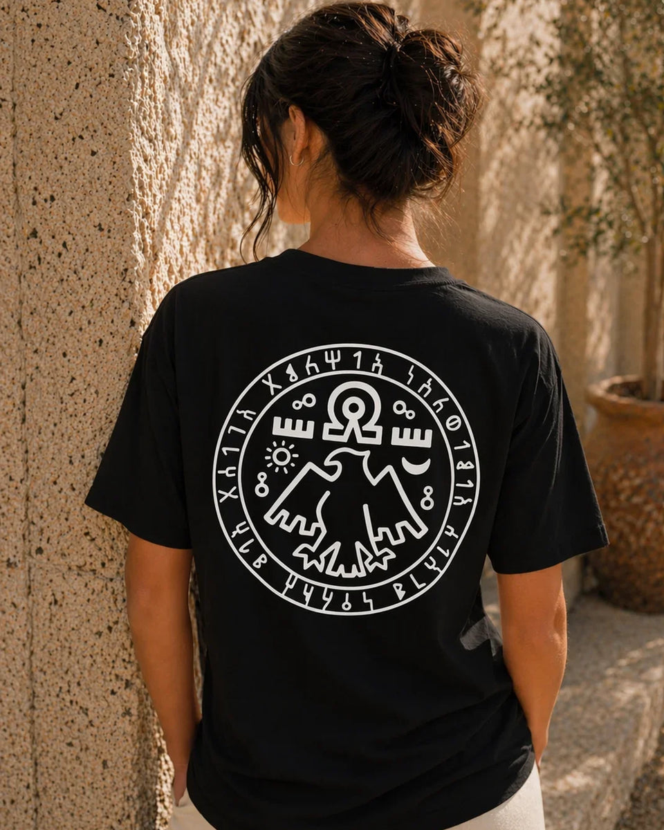 Person wearing a black t-shirt with a white symbol on the back, standing against a textured wall.