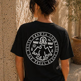 Person wearing a black t-shirt with a white symbol on the back, standing against a textured wall.