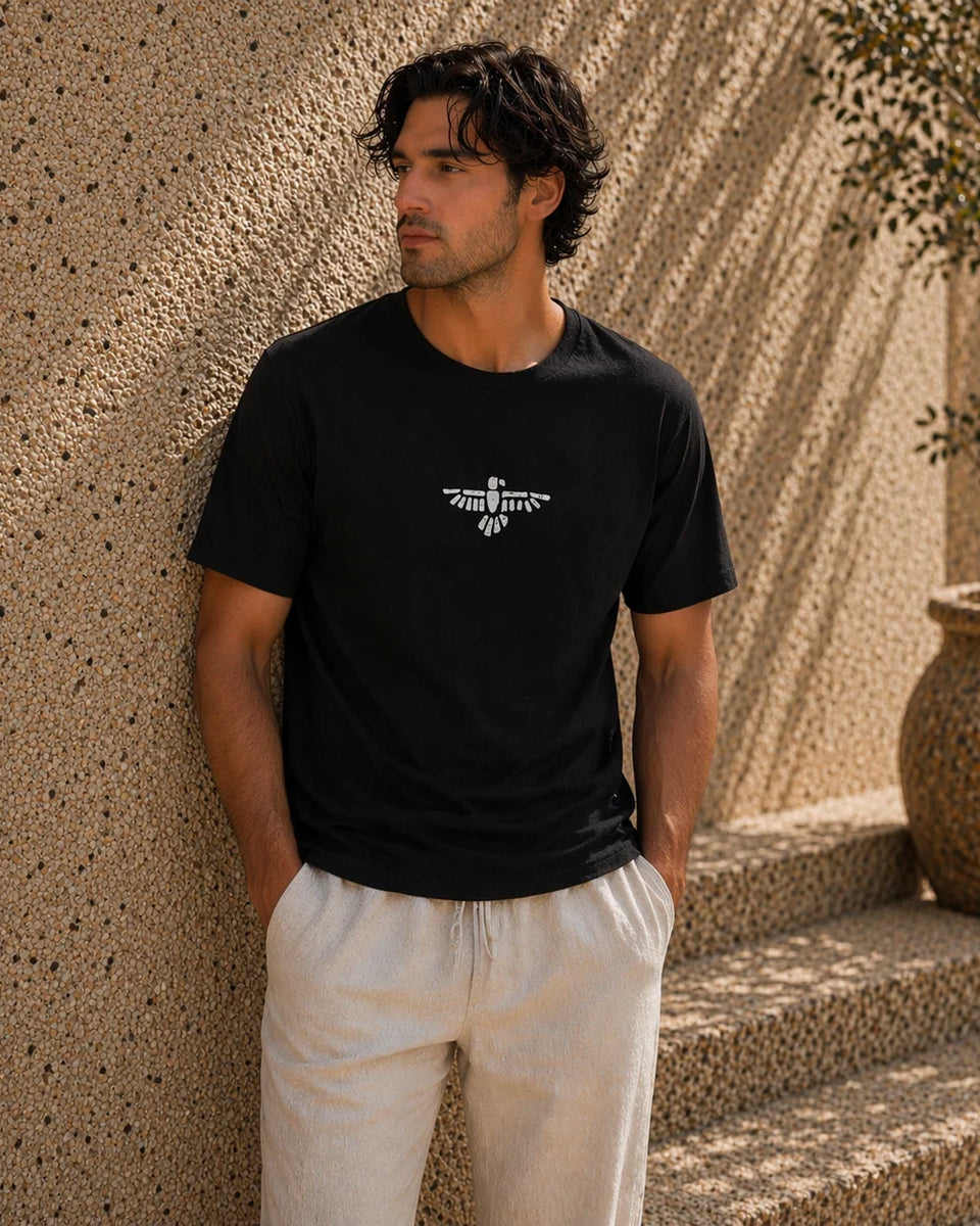 Man wearing a black t-shirt with a logo standing against a textured wall.