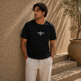 Man wearing a black t-shirt with a logo standing against a textured wall.