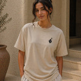Woman wearing a beige t-shirt with a black logo, standing outdoors.