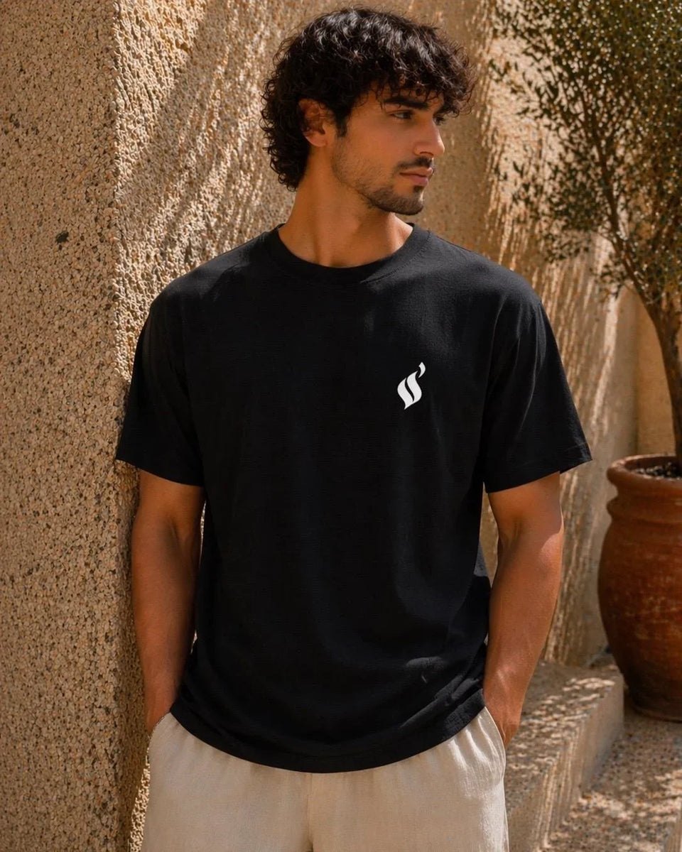 Man wearing a black t-shirt with a white logo against a textured wall.