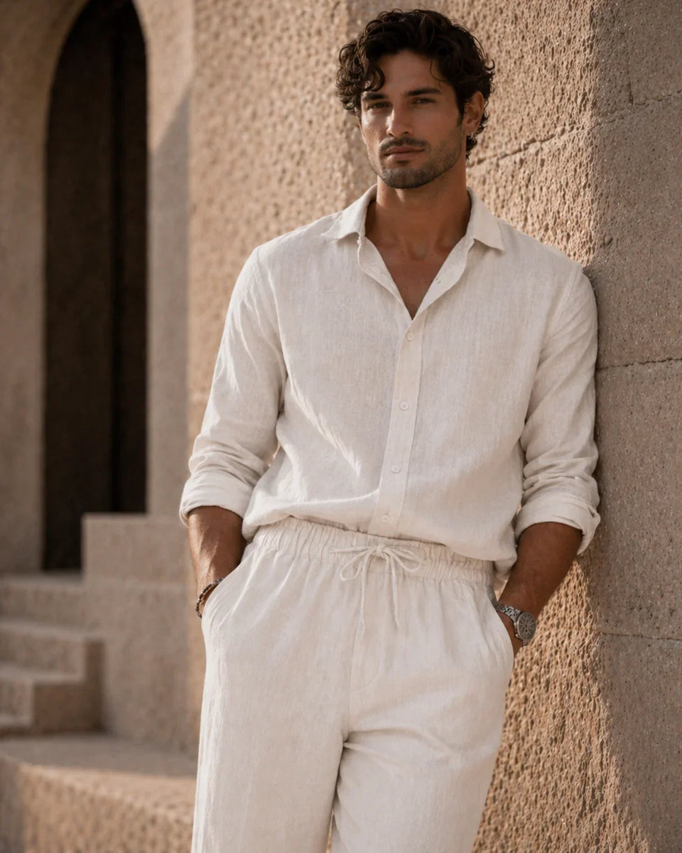 Man wearing a light-colored shirt and pants leaning against a stone wall.