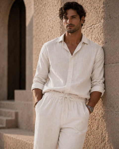 Man wearing a light-colored shirt and pants leaning against a stone wall.