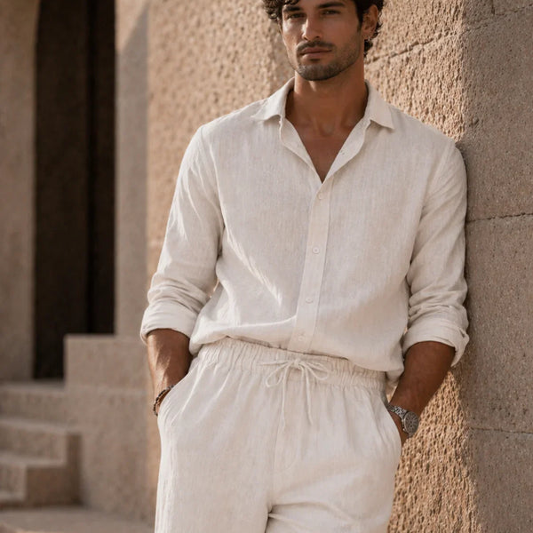 Man wearing a light-colored shirt and pants leaning against a stone wall.