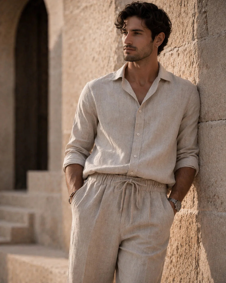 Man wearing a beige linen shirt and pants leaning against a stone wall.