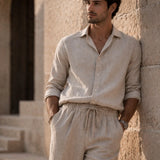 Man wearing a beige linen shirt and pants leaning against a stone wall.