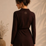 Woman wearing a dark brown dress with a subtle design on the back against a beige wall.