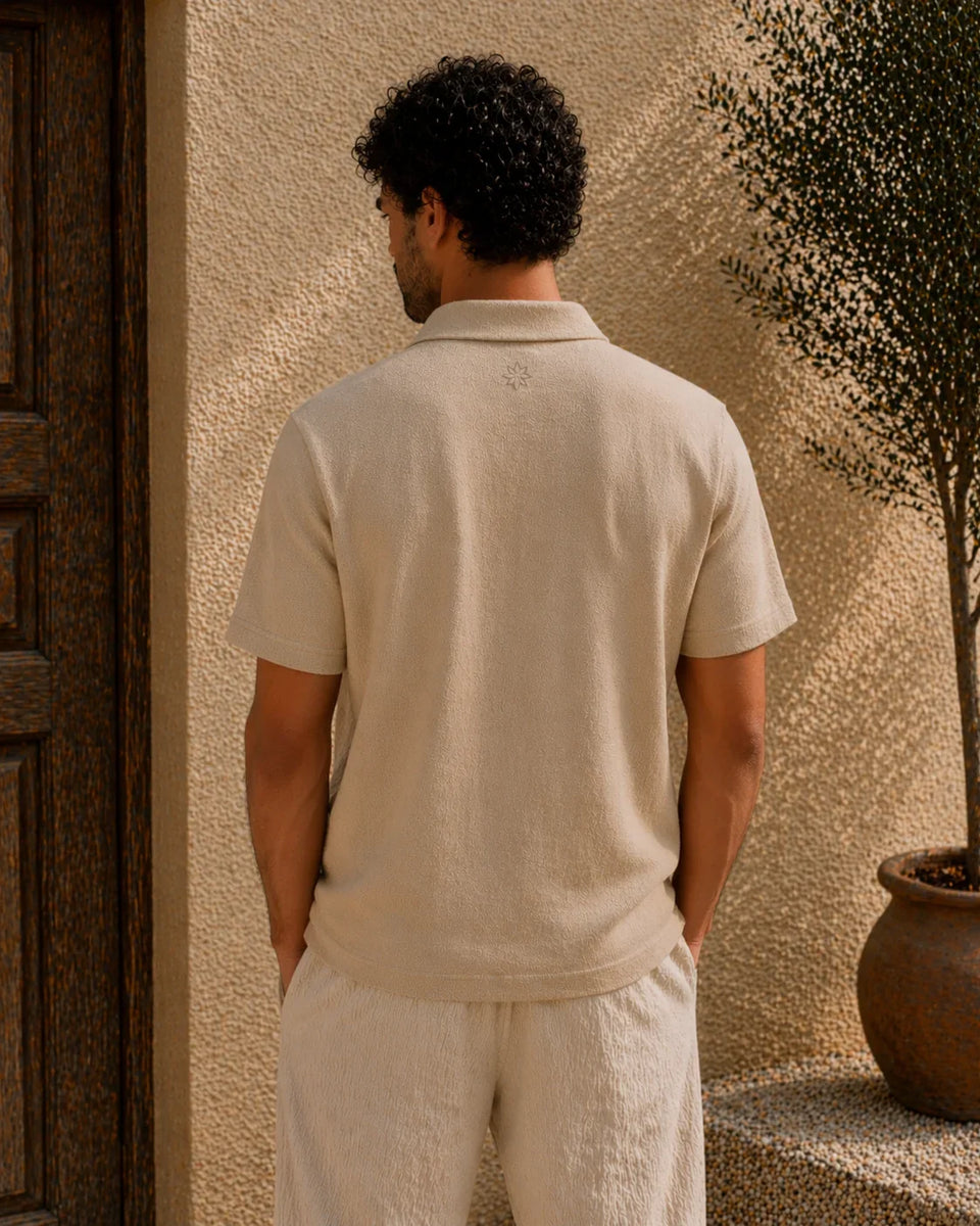 Man wearing a beige polo shirt and pants standing against a textured wall.