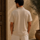 Man wearing a beige polo shirt and pants standing against a textured wall.