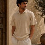 Man wearing a beige polo shirt and pants standing against a textured wall.