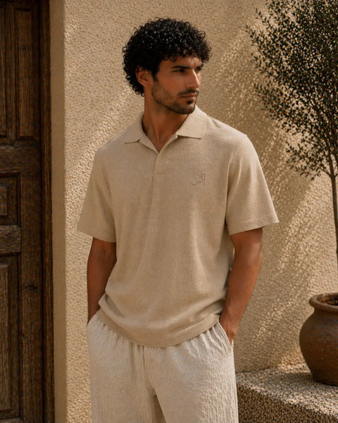 Man wearing a beige polo shirt and pants standing against a textured wall.