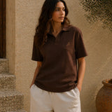 Woman wearing a brown polo shirt with a logo, standing against a textured wall.