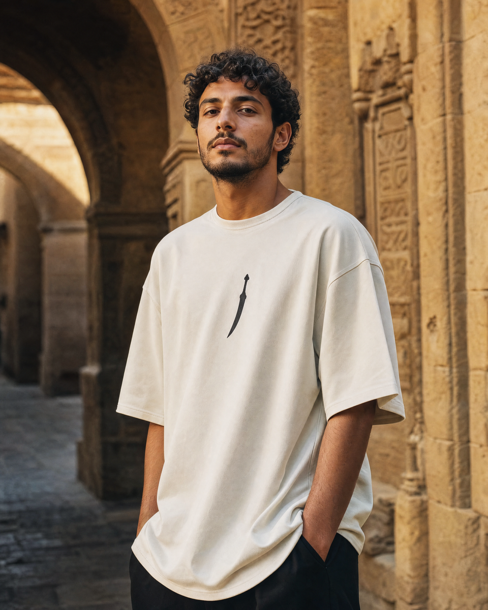 Man wearing QADIM Jambiya Oversize T-Shirt in Beige, front view with small dagger icon on chest