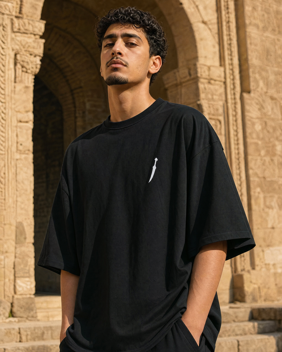 Man wearing QADIM Jambiya Oversize T-Shirt in Black, front view with small white dagger icon on chest