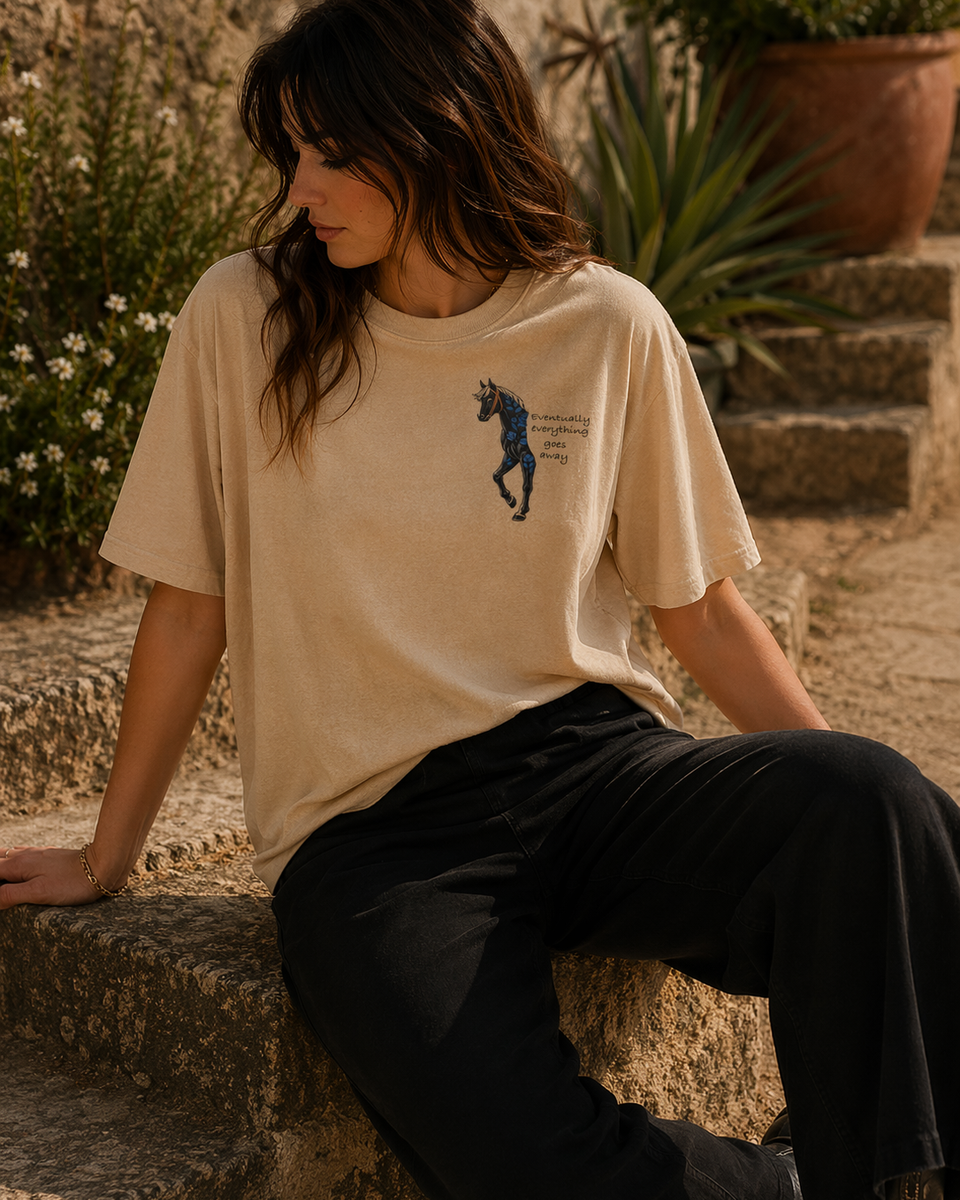 Woman wearing a beige t-shirt with a graphic design, sitting on stone steps outdoors.