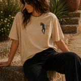 Woman wearing a beige t-shirt with a graphic design, sitting on stone steps outdoors.
