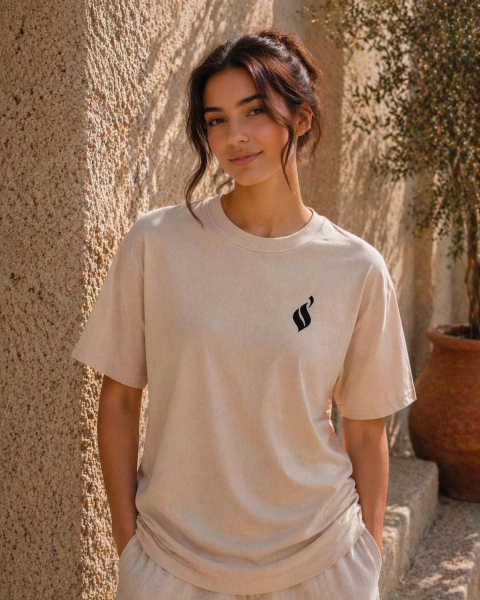 Woman wearing a beige t-shirt with a black logo against a textured wall.