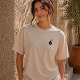 Woman wearing a beige t-shirt with a black logo against a textured wall.
