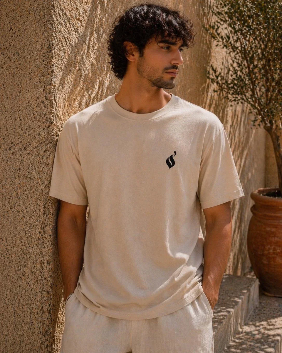 Man wearing a beige t-shirt with a black logo against a textured wall.