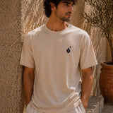 Man wearing a beige t-shirt with a black logo against a textured wall.