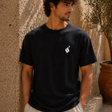 Man wearing a black t-shirt with a white logo against a textured wall.