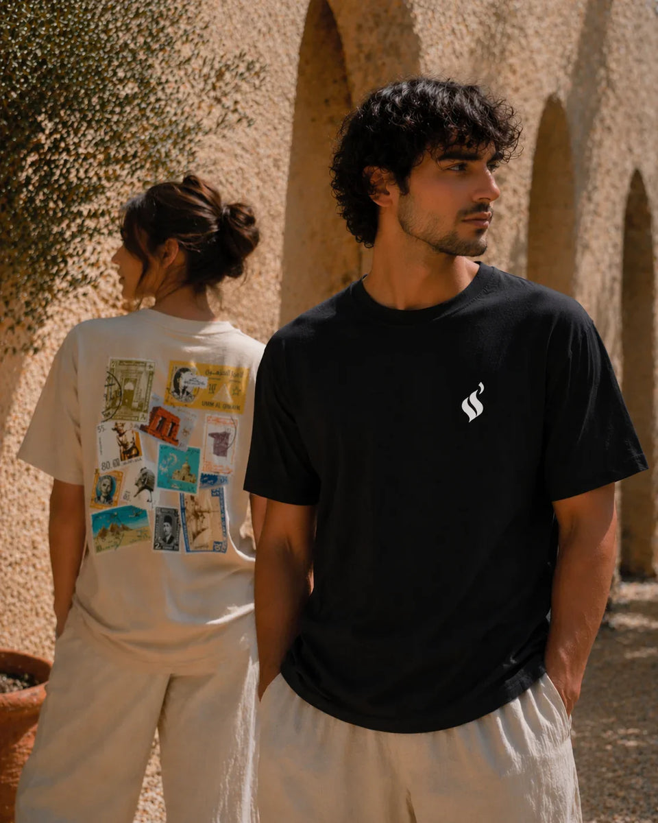 Two people standing outdoors with a man wearing a black t-shirt with a logo and a woman wearing a white t-shirt with a graphic design.
