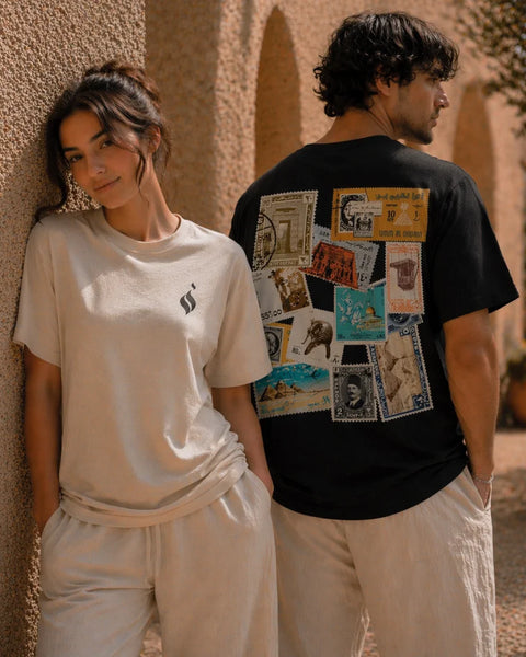 Two people standing outdoors, one wearing a white t-shirt with a logo and the other wearing a black t-shirt with vintage stamp designs.