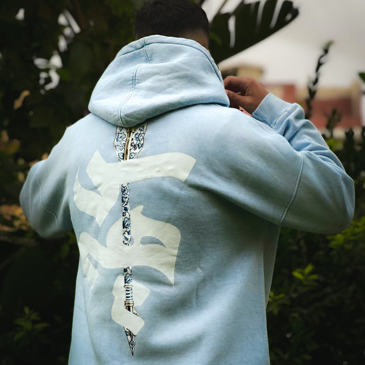 QADIM Sahm washed hoodie in baby blue, back view in a garden | هودي سهم مغسول بيبي بلو في جنينة