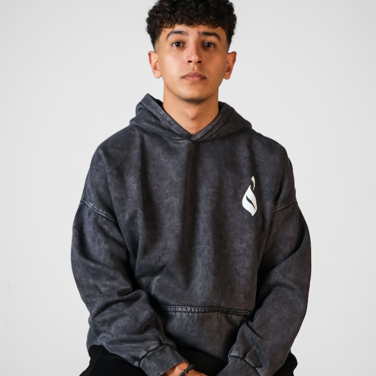 QADIM Sahm washed hoodie in black, front emblem, seated model | هودي سهم مغسول أسود من قدّام